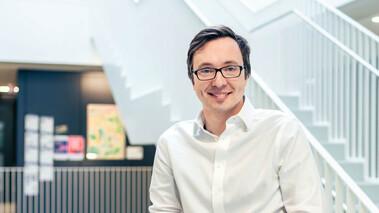 Porträt von Alexey Chernikov in weißem Hemd, lächelnd, vor einer modernen Treppe mit hellem Hintergrund.